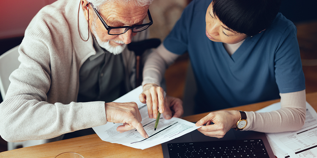 A healthcare professional assists an elderly man with paperwork, pointing at information on a form.